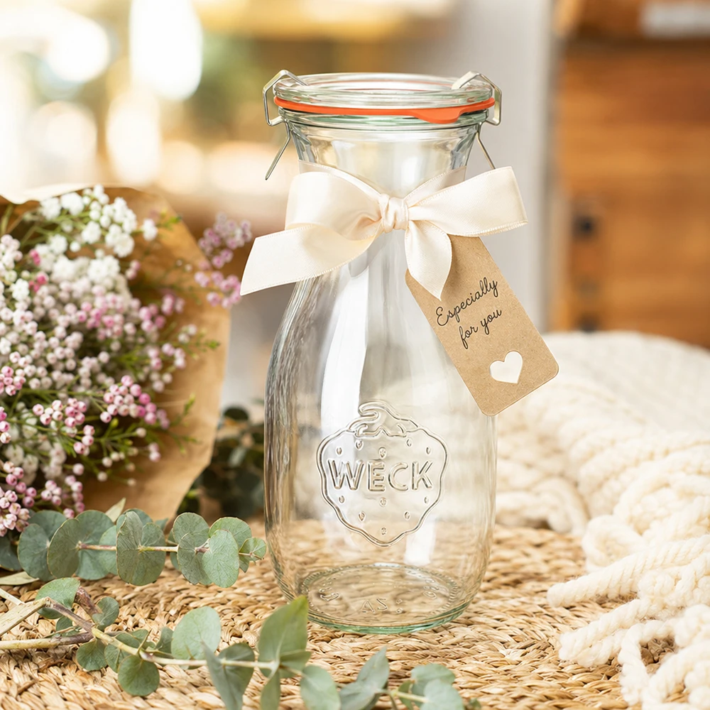 WECK 530 ml juiceflaska RR60 dekorerad med ett vitt band och en presentetikett placerad mellan blommor