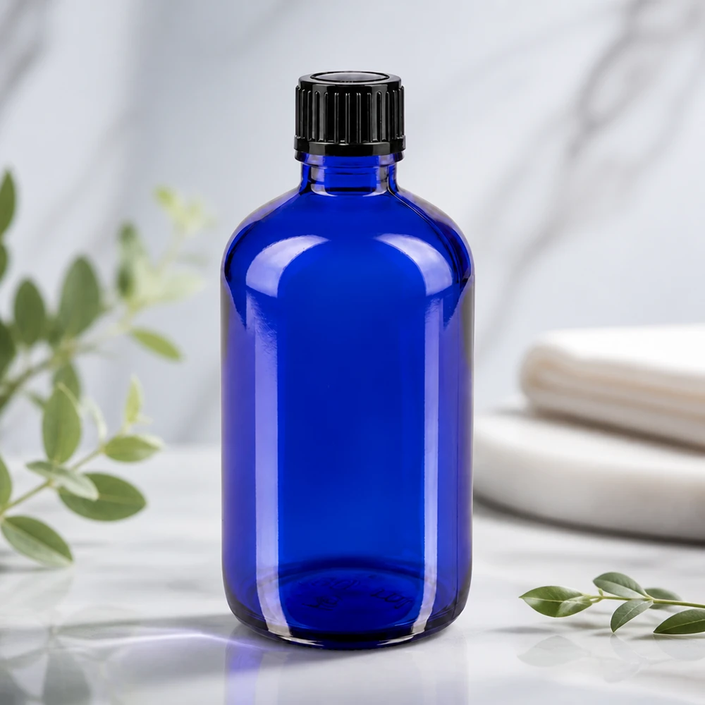 Apoteksflaska blå 100ml dropplock 1mm svart standard Blå glasflaska på 100 ml med räfflad svart kork på en vit marmoryta med eukalyptusgrenar