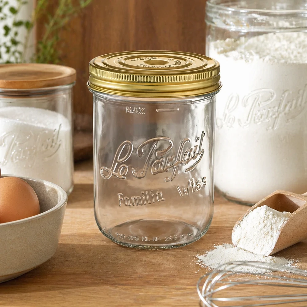 Le Parfait® Familia Wiss 1 kg Ø100 mm burk 1020 ml Familia Wiss burk med guldlock på träbord bredvid bakingredienser och andra konserveringsburkar