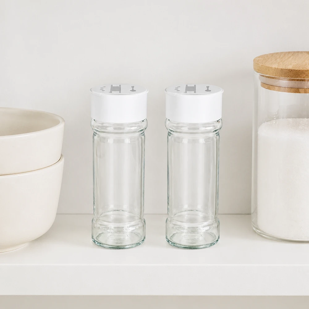 Salt- och kryddburk 100 ml med dubbel ströare, vit Två kryddburkar à 100 ml med vita lock med dubbla ströare, placerade bredvid varandra på en vit hylla